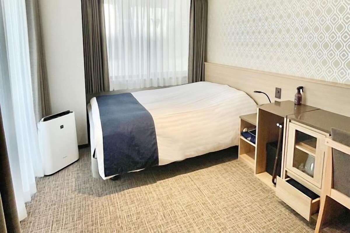 Habitació d'hotel amb un llit, un escriptori amb un ordinador, un petit armari i una finestra amb cortines.