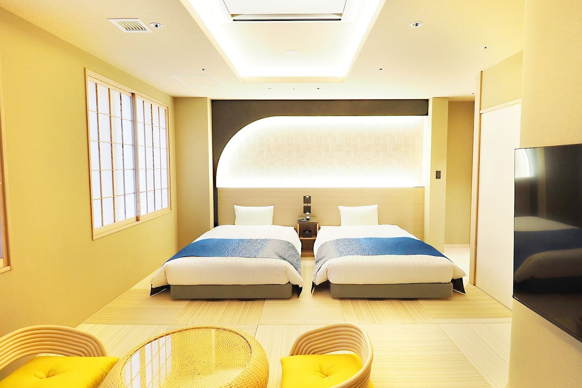 Habitación de hotel con dos camas, televisor y zona de estar con cojines amarillos.