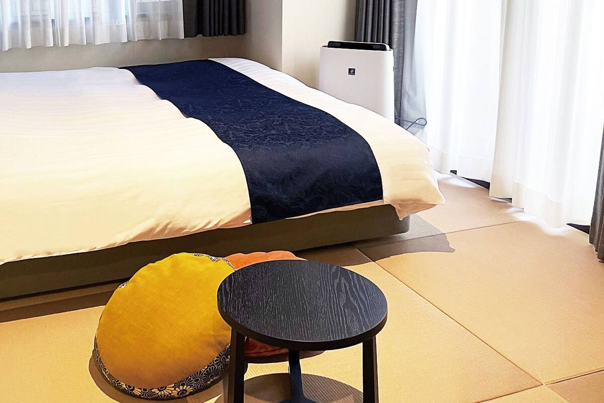 Una cama bien hecha con una colcha blanca y azul oscuro, una mesita redonda con tapa oscura y un almohadón amarillo.