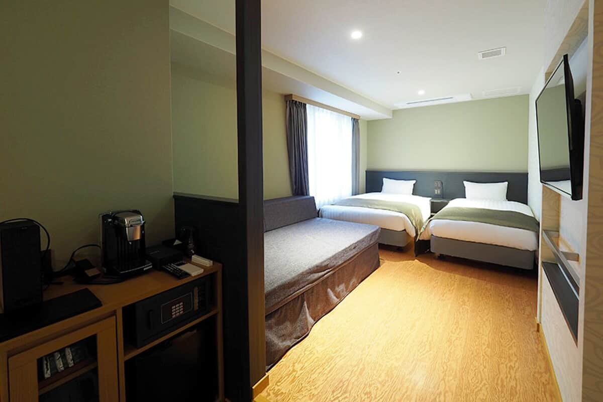 Habitación de hotel con dos camas, un escritorio con cafetera, televisor y ventana con cortinas.
