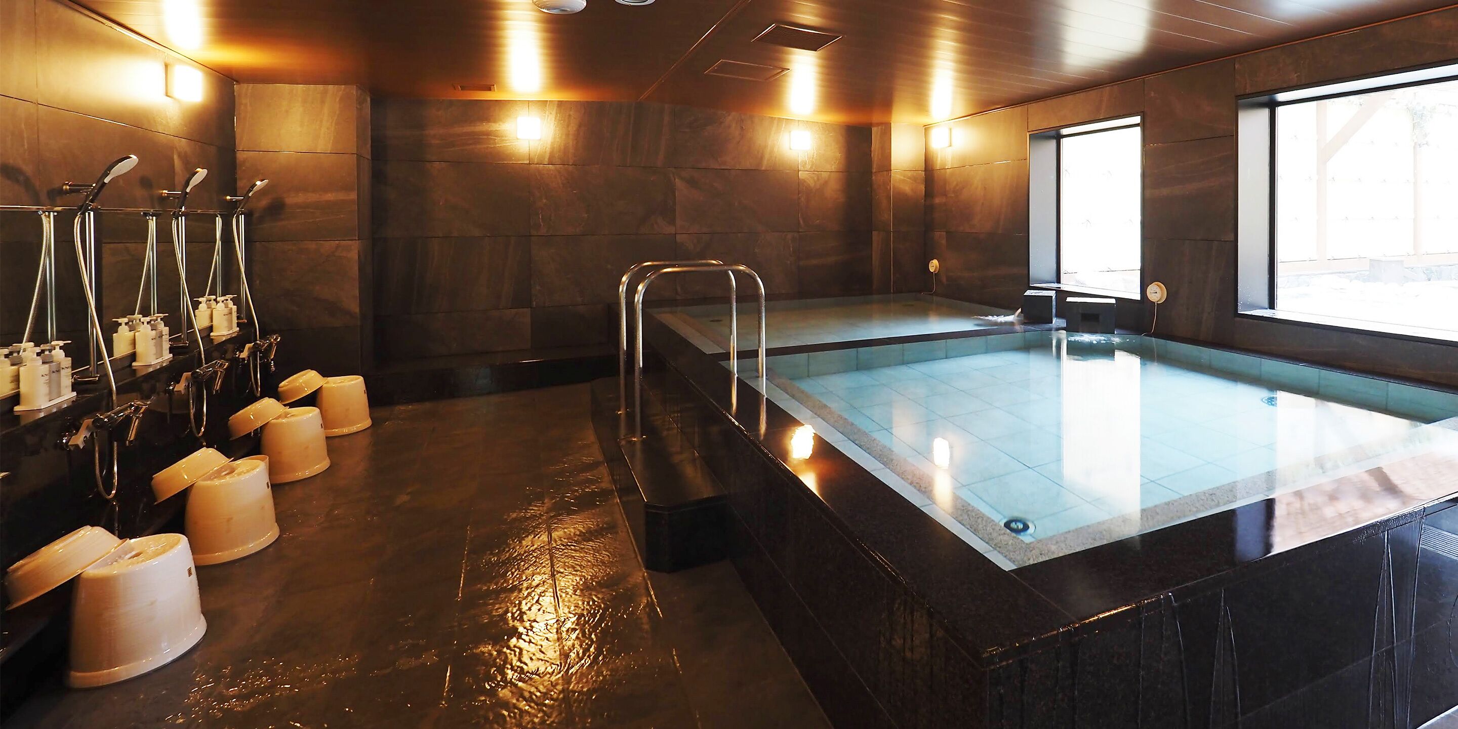 A modern indoor pool with a glass barrier, multiple showerheads, and a sunlit room with stone walls.
