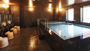 A modern indoor pool with a glass barrier, multiple showerheads, and a sunlit room with stone walls.