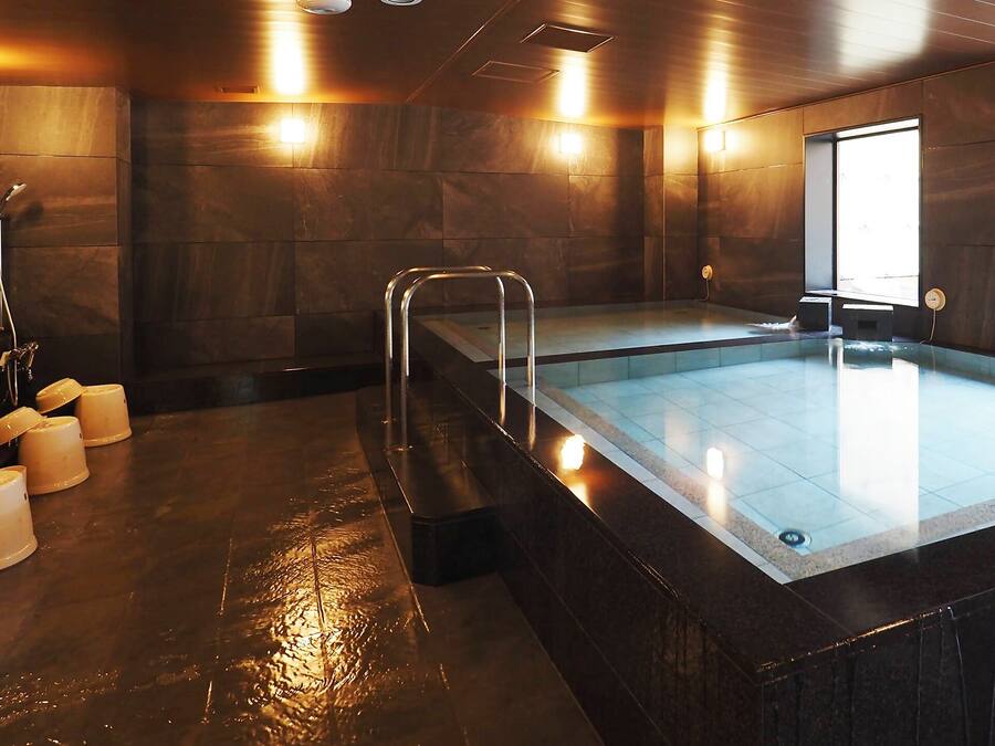 A modern indoor pool with a glass barrier, multiple showerheads, and a sunlit room with stone walls.