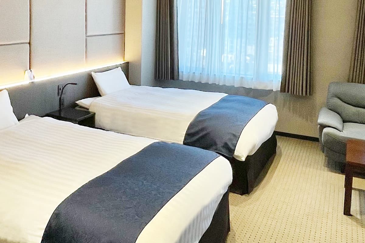 Habitación de hotel con dos camas, una mesita de noche, un sofá y una ventana con cortinas.
