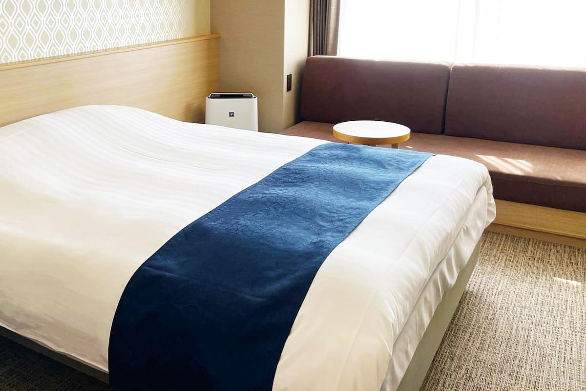 Una habitación de hotel con cama, mesita de noche pequeña, sillón y sofá, además de una ventana.