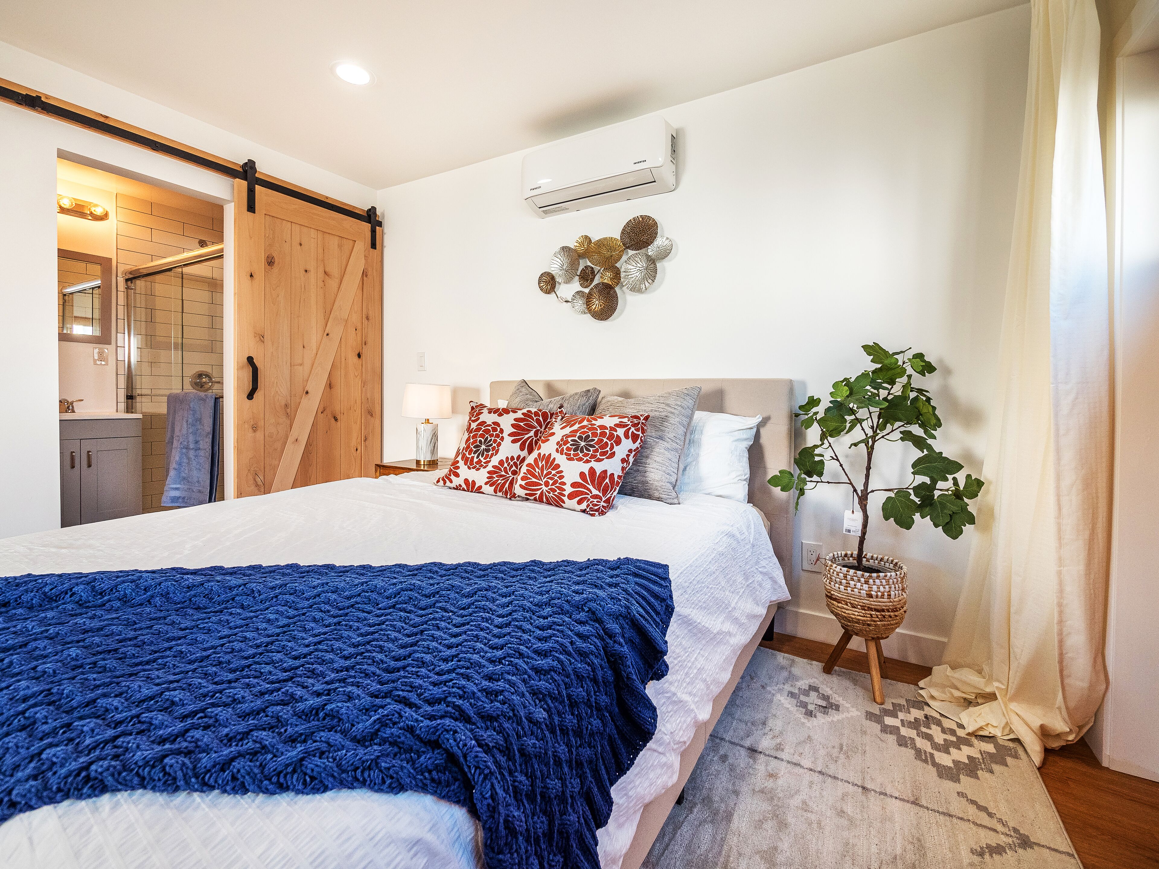 Un dormitorio con cama, mesita de noche, aire acondicionado de pared, planta en maceta y puerta corrediza que da a un baño.