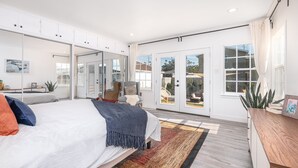 A modern bedroom with a large bed, a seating area with a chair and table, and a sliding glass door leading to a patio.
