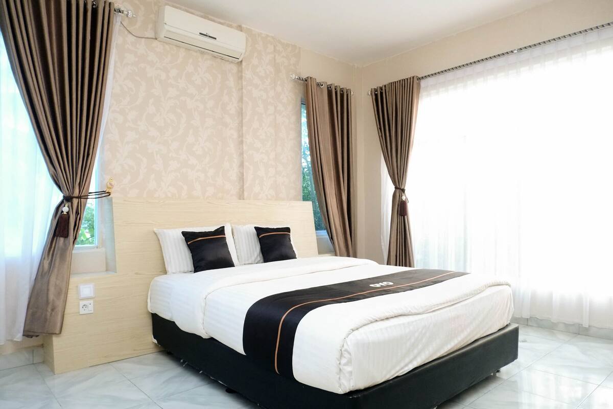 Une chambre d’hôtel moderne avec un grand lit, des murs aux motifs beiges et une fenêtre avec des rideaux.