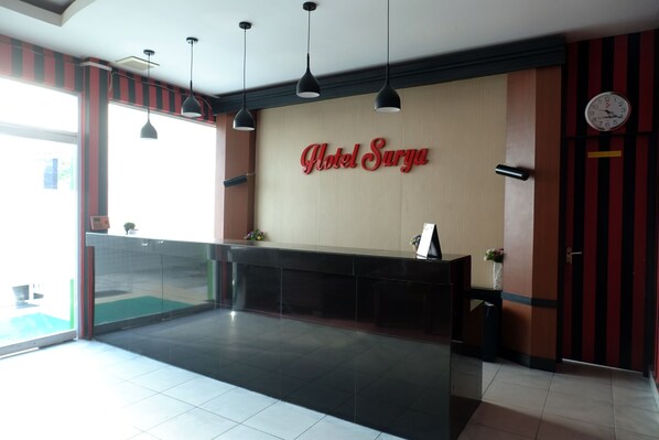 A hotel reception area with a modern design, featuring a reception desk, a clock, and a large sign that reads "Hotel Surya".