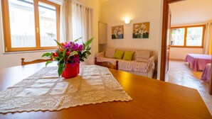 A cozy dining area with a wooden table, a lace tablecloth, and a vase of flowers. There is a sofa with a floral cushion and a painting on the wall.