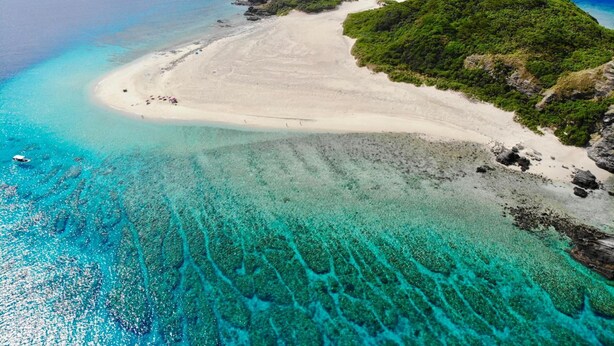 Vista aérea de una playa con aguas turquesas cristalinas, una orilla de arena y colinas verdes exuberantes al fondo.