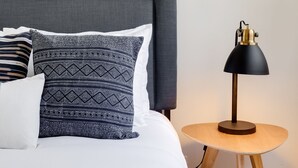 Cama bem arrumada com almofadas estampadas e uma pequena mesa redonda com abajur ao lado.