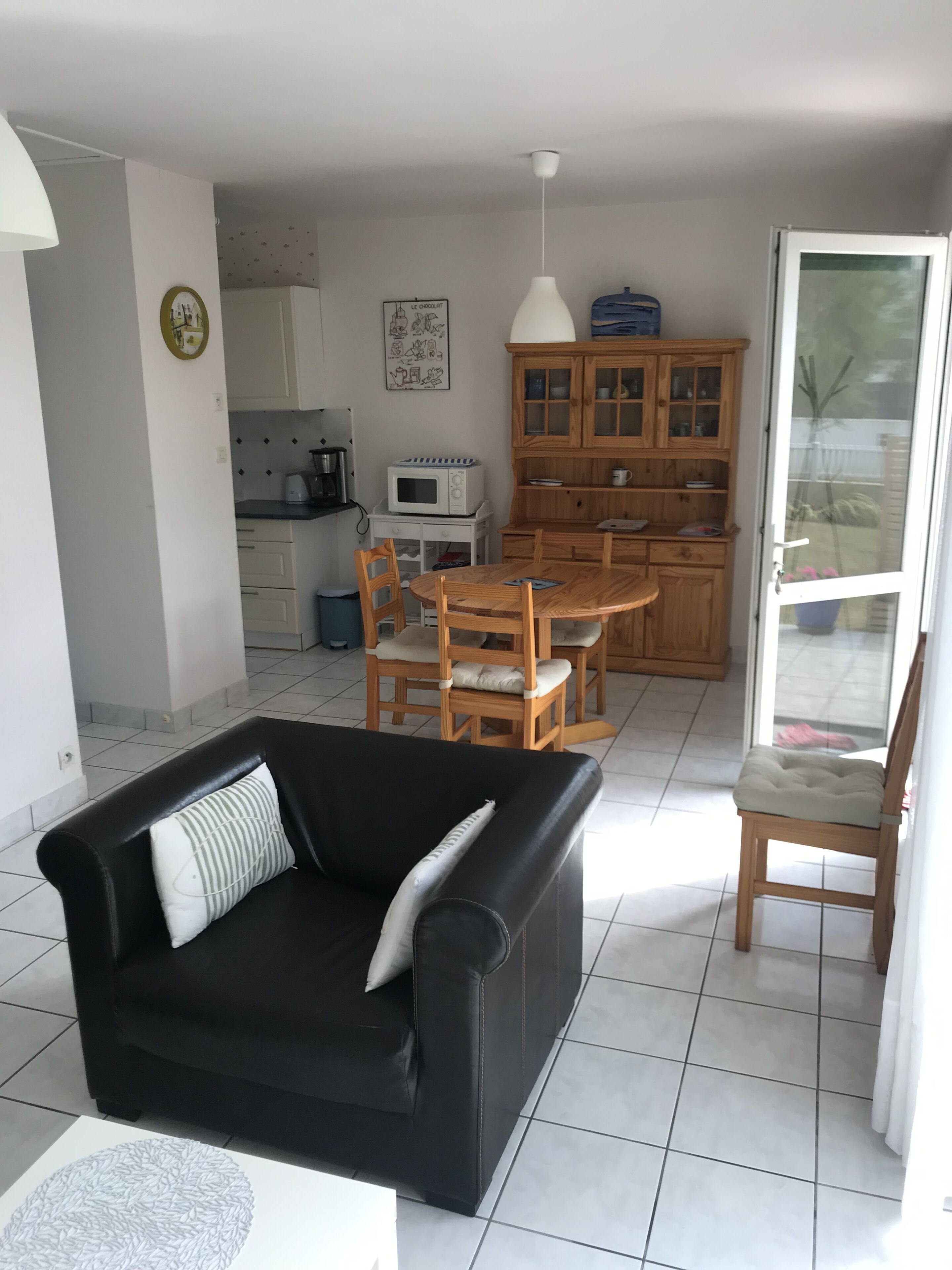 A compact living space with a black sofa, wooden dining table, and chairs, a kitchen area with cabinets and appliances, and a sliding glass door leading to a garden.