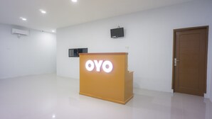 Uma área de recepção com balcão de madeira exibindo o logotipo "OYO", uma TV de tela plana na parede e uma porta de madeira fechada.
