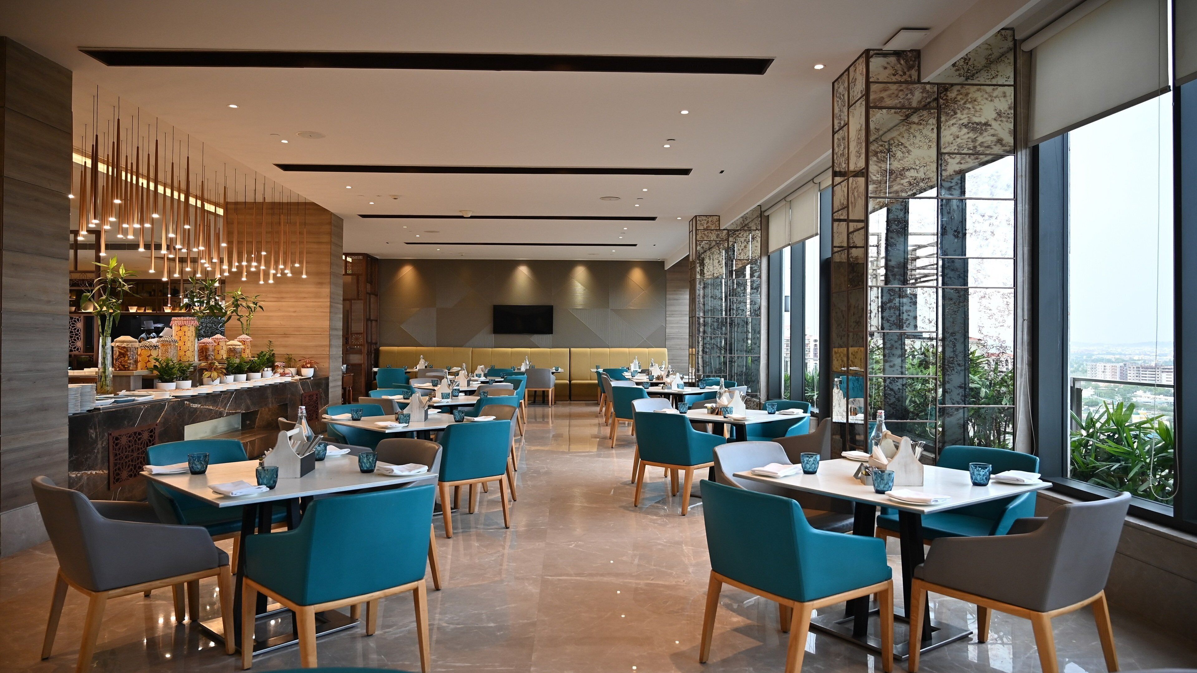 A modern restaurant with teal chairs and tables, a bar area with jars, and large windows offering a city view.