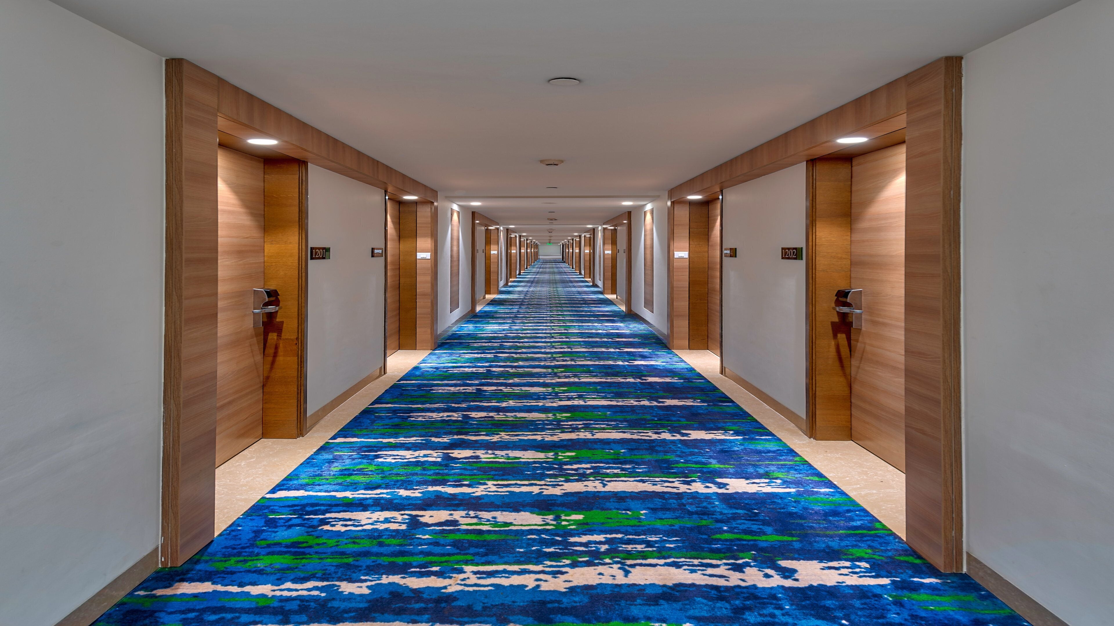 A long hallway with a blue patterned carpet, wooden paneling on the walls, and doors with room numbers.