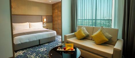 A hotel room with a bed, a sofa, a coffee table with fruits and a card, and a window with curtains.