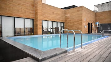 A swimming pool with a wooden deck, metal railings, and a modern building with large windows in the background.