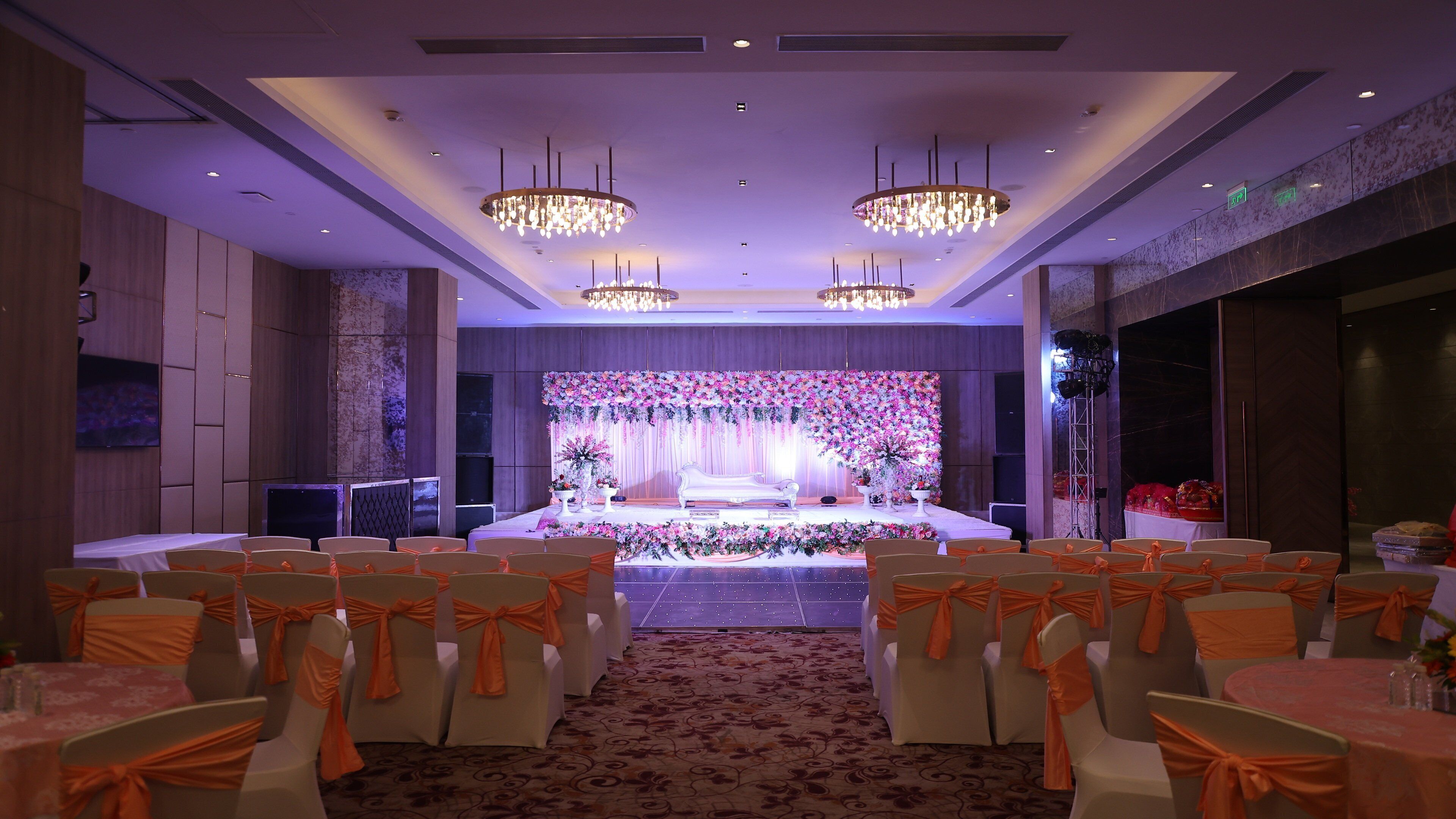A banquet hall with a stage, rows of tables with chairs, and chandeliers.