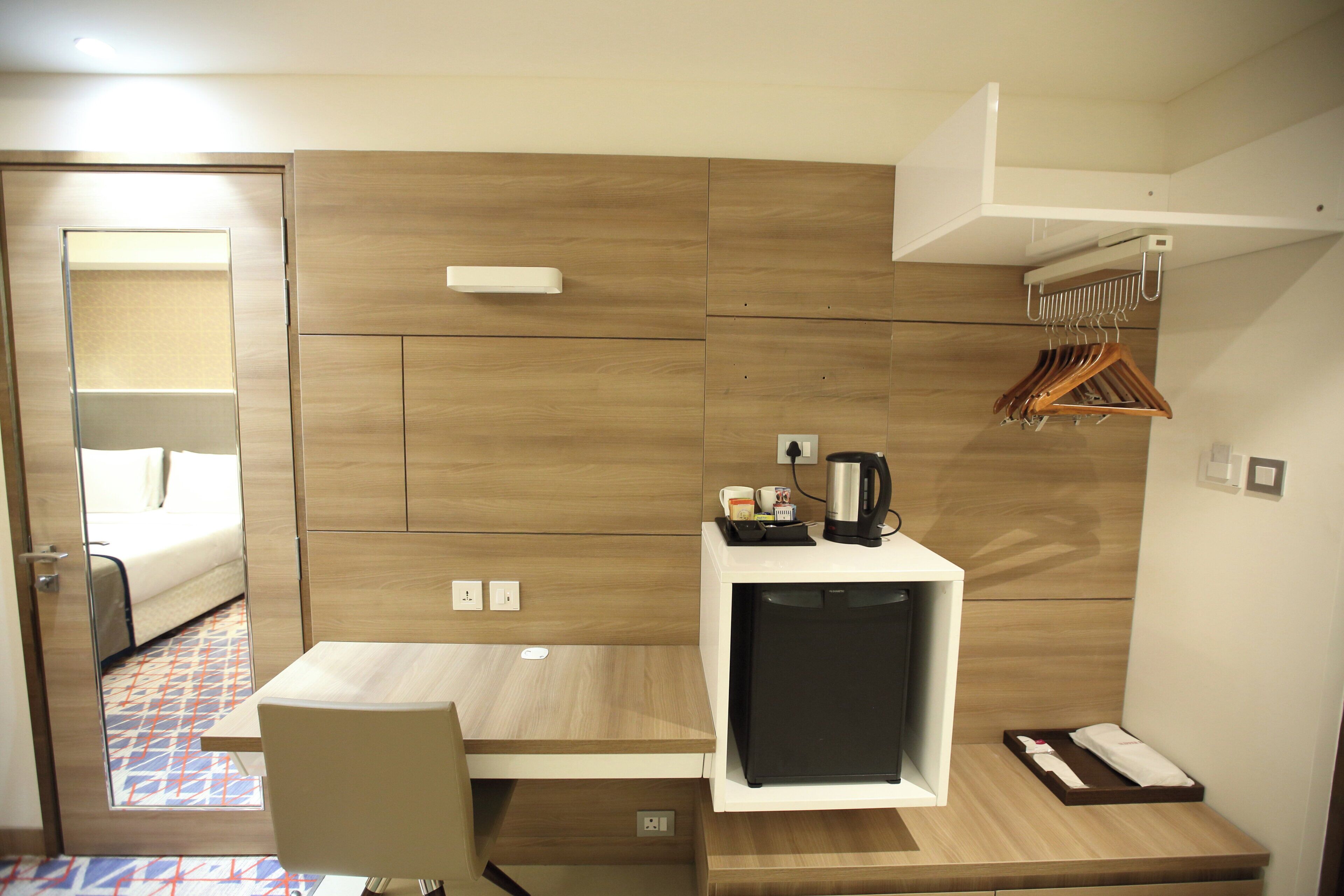 A compact hotel room with a bed, a small desk, a coffee maker, and a wardrobe with hanging clothes.