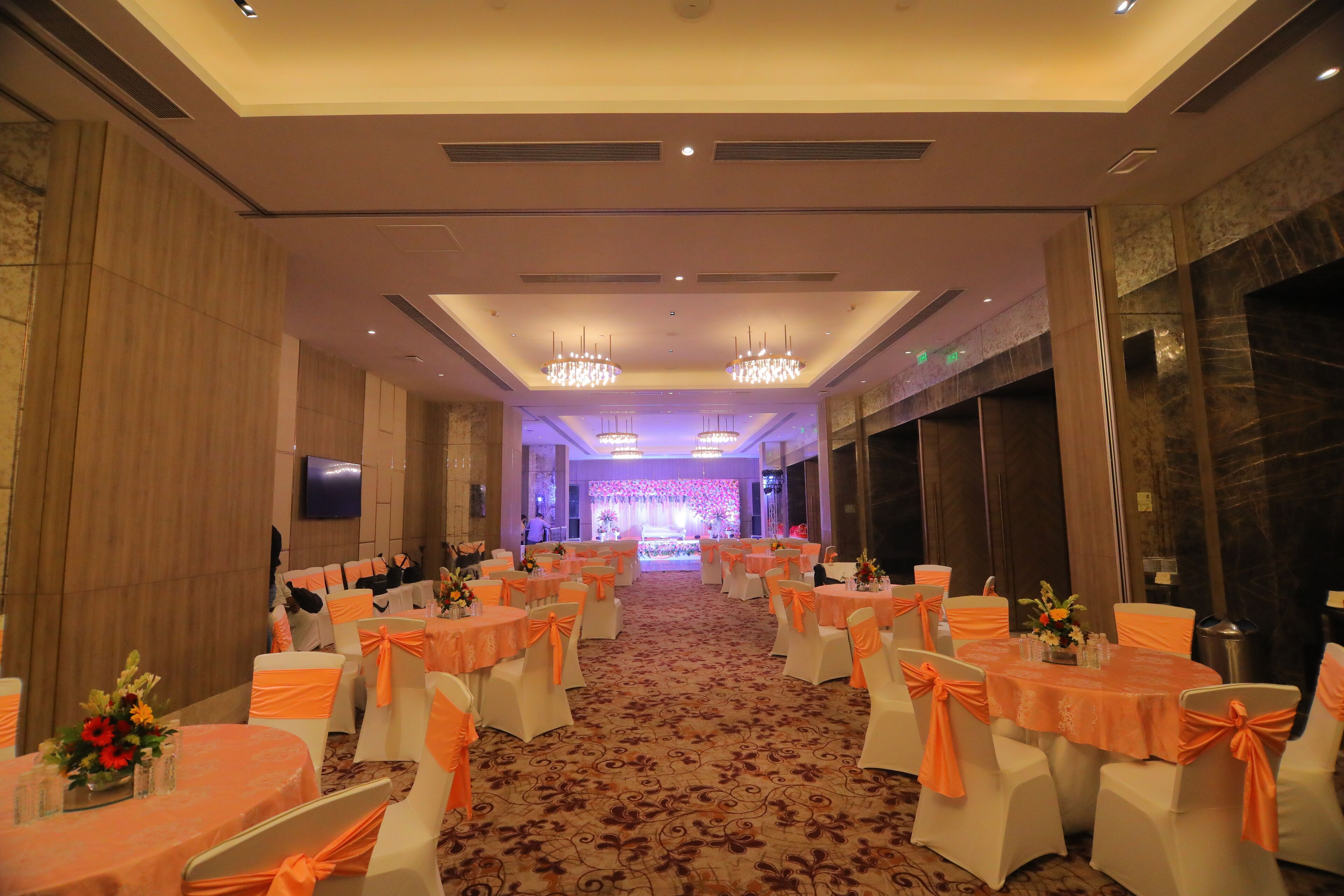 A banquet hall with round tables, white chairs with orange sashes, and chandeliers.