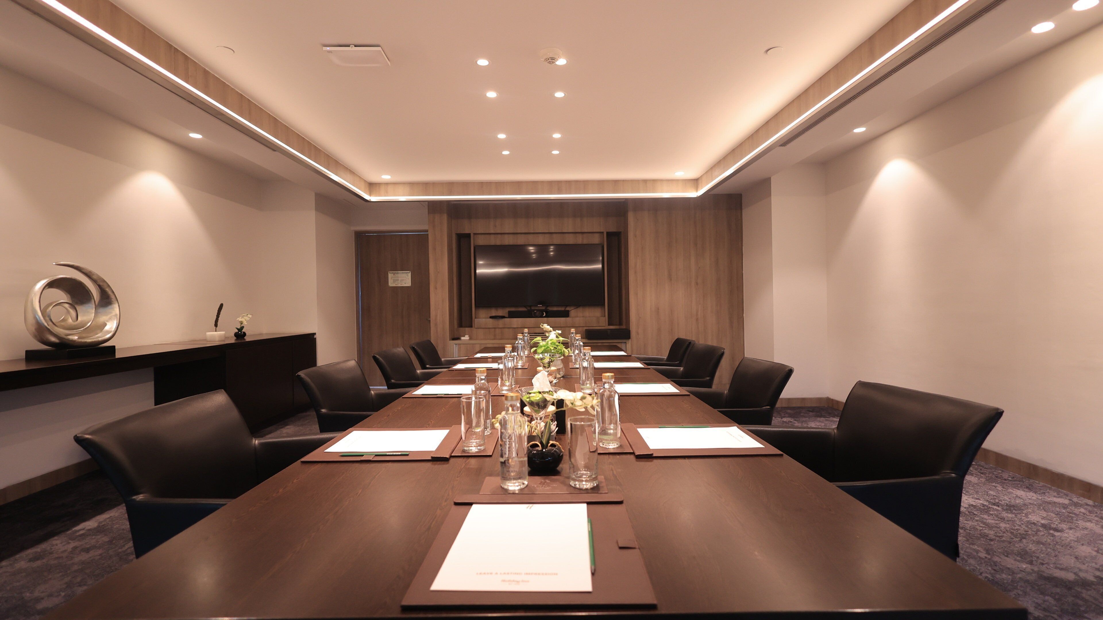 A conference room with a long table, chairs, a TV screen, and a decorative vase.
