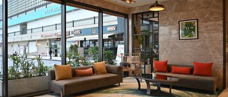 A modern lobby with a grey sofa, red cushions, a coffee table, and large windows offering a view of the street and buildings.