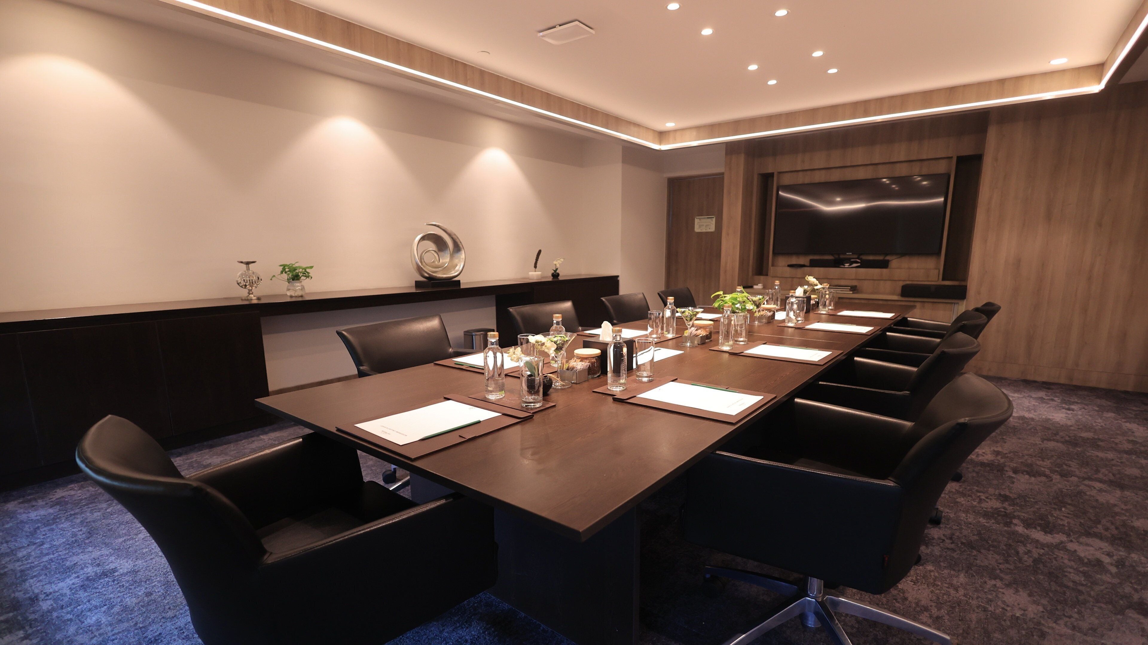A conference room with a long table, black chairs, and a large screen.