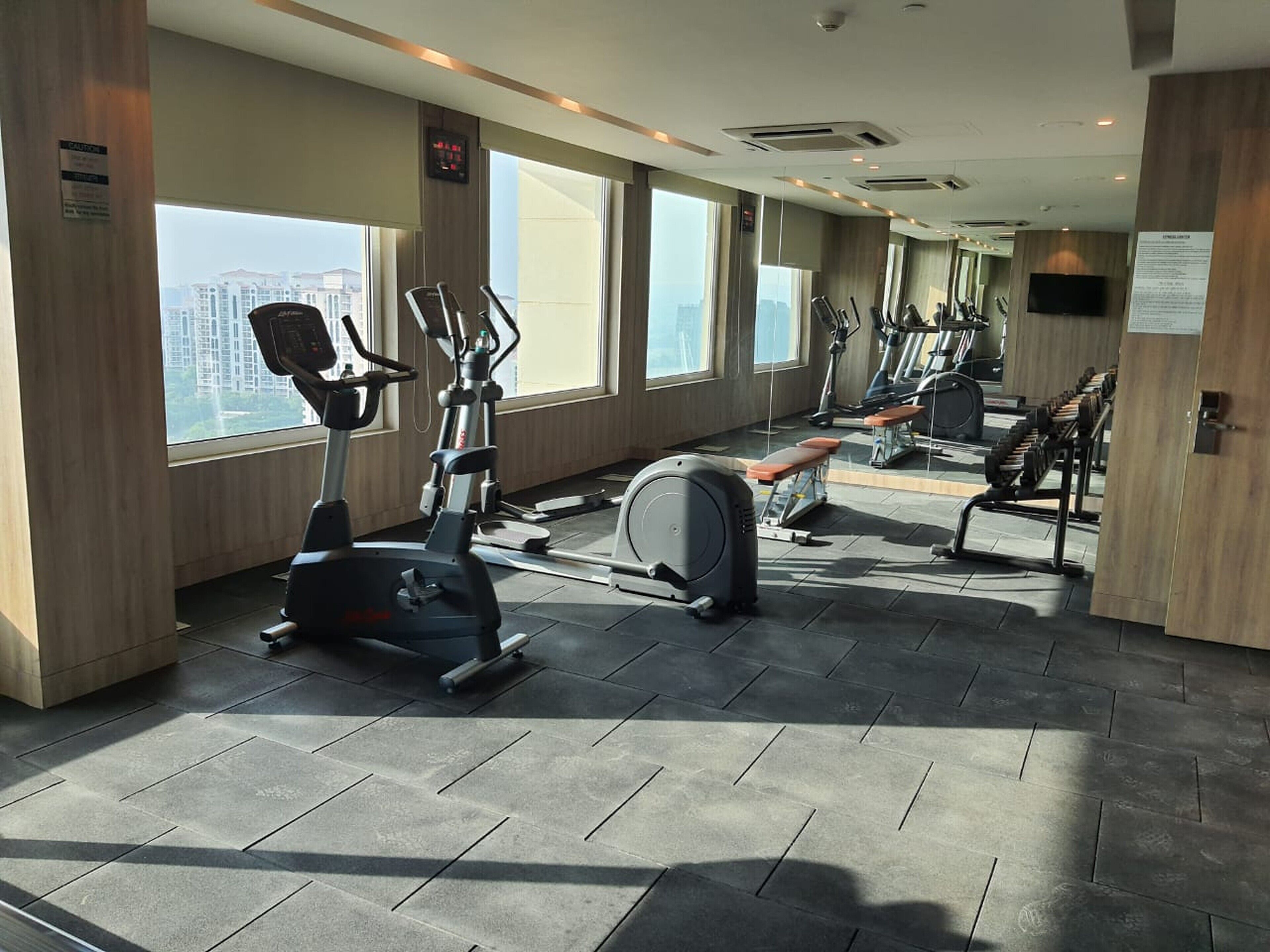 A modern gym with various cardio machines, a bench, and a large window offering a city view.