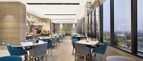 A modern restaurant with high ceilings, large windows offering a city view, and a variety of seating options.