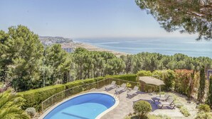 Une piscine à bord incurvé, entourée d’une terrasse dallée et d’une rambarde en métal. On y trouve des chaises longues et une petite table avec un parasol. La piscine est située dans un espace aménagé avec des arbres et des arbustes.