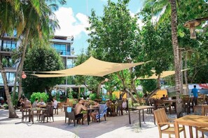 An outdoor dining area with tables and chairs, shaded by a large canopy, surrounded by palm trees and greenery. - Seashore Beach Inn (Maafushi)