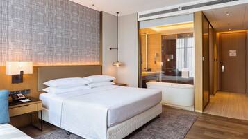 A modern hotel room with a large bed, bedside table, and a bathroom with a bathtub and glass shower enclosure.