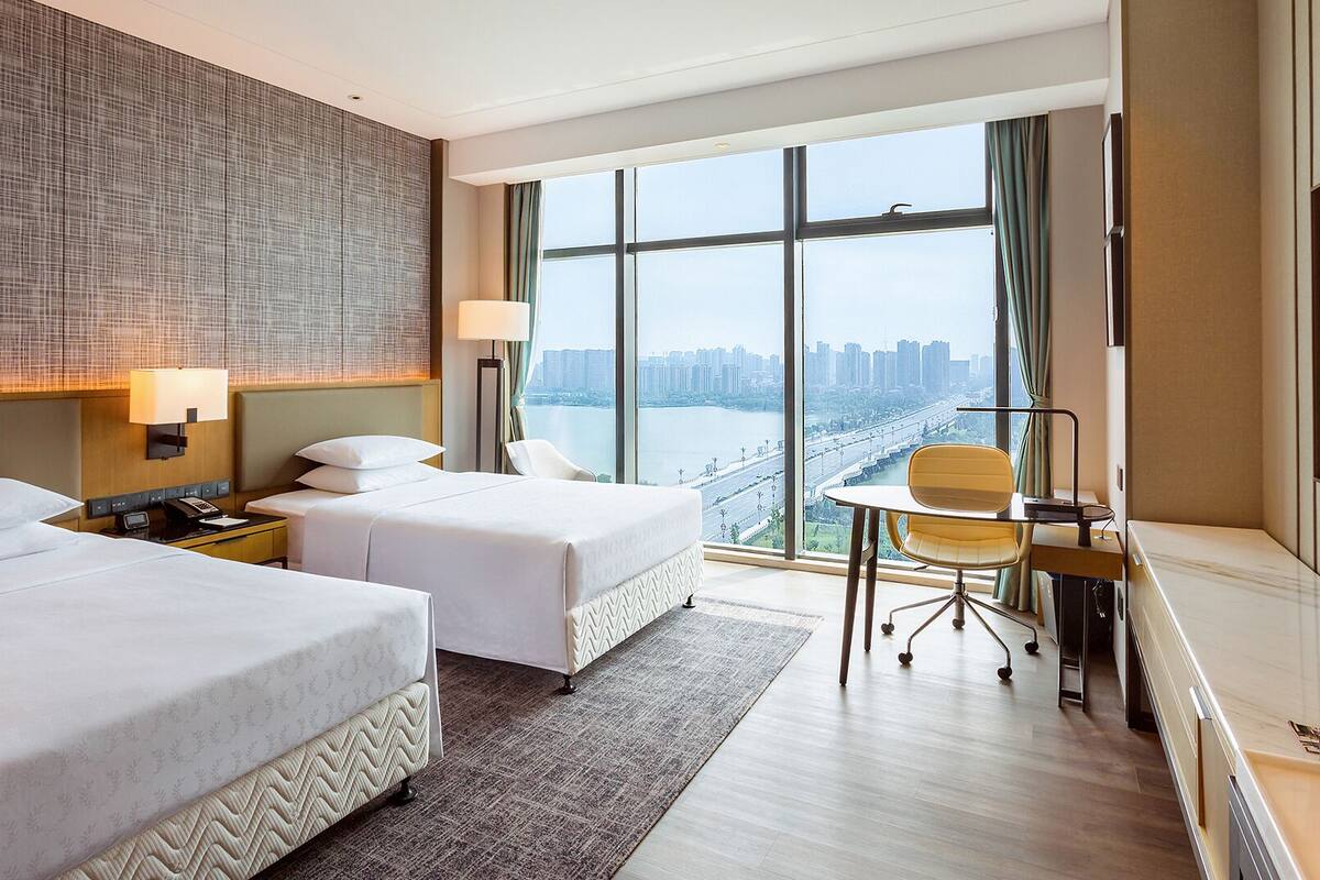 Ein Hotelzimmer mit zwei Betten, einem Schreibtisch mit Stuhl, einem Fernseher und einem großen Fenster mit Stadtblick.