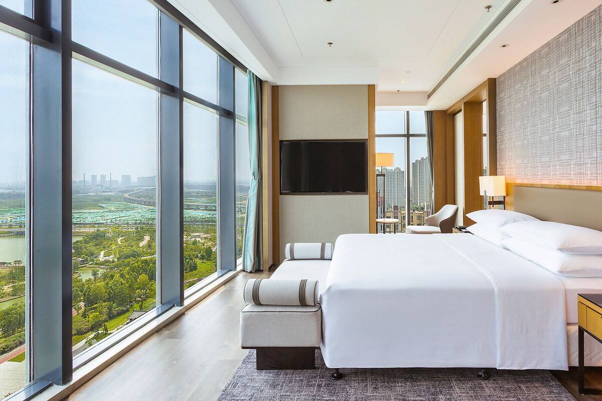 Ein modernes Hotelzimmer mit einem großen Bett, einem Schreibtisch und einem Fernseher. Große Fenster bieten Blick auf die Stadt.