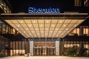 Entrada de un hotel Sheraton con diseño moderno, que presenta un gran letrero iluminado, un amplio vestíbulo y una fachada bien iluminada.