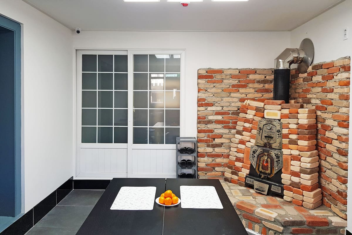 Una habitación con una mesa negra, una pizarra y un plato de naranjas. Se observa una pared de ladrillo con una chimenea y una ventana con vidrio esmerilado.