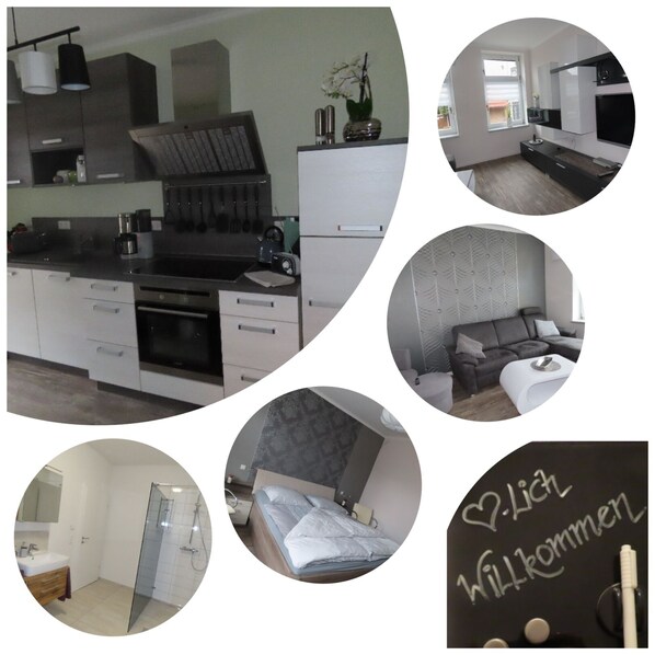 A modern kitchen with a built-in oven, a dining area with a sofa, a bathroom with a glass shower, and a bedroom with a bed and a bedside table.