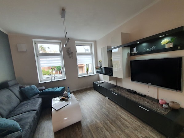 A modern living room with a grey sofa, a white coffee table, and a large flat-screen TV mounted on the wall.