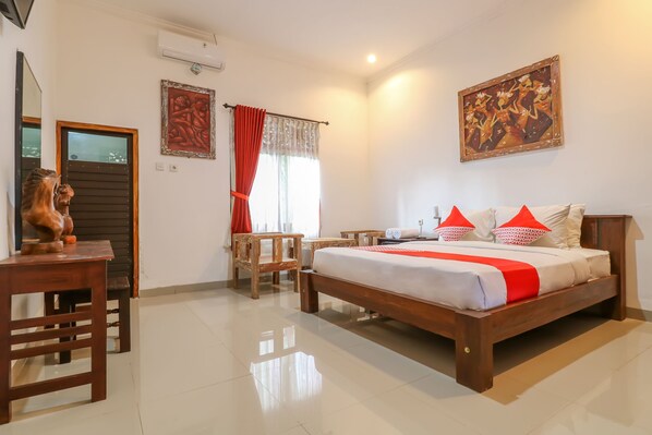 A bedroom with a wooden bed, a desk, a chair, and a small table. There is a window with curtains, a painting on the wall, and a door. - OYO 2607 Men Bebas Residence (Ubud)