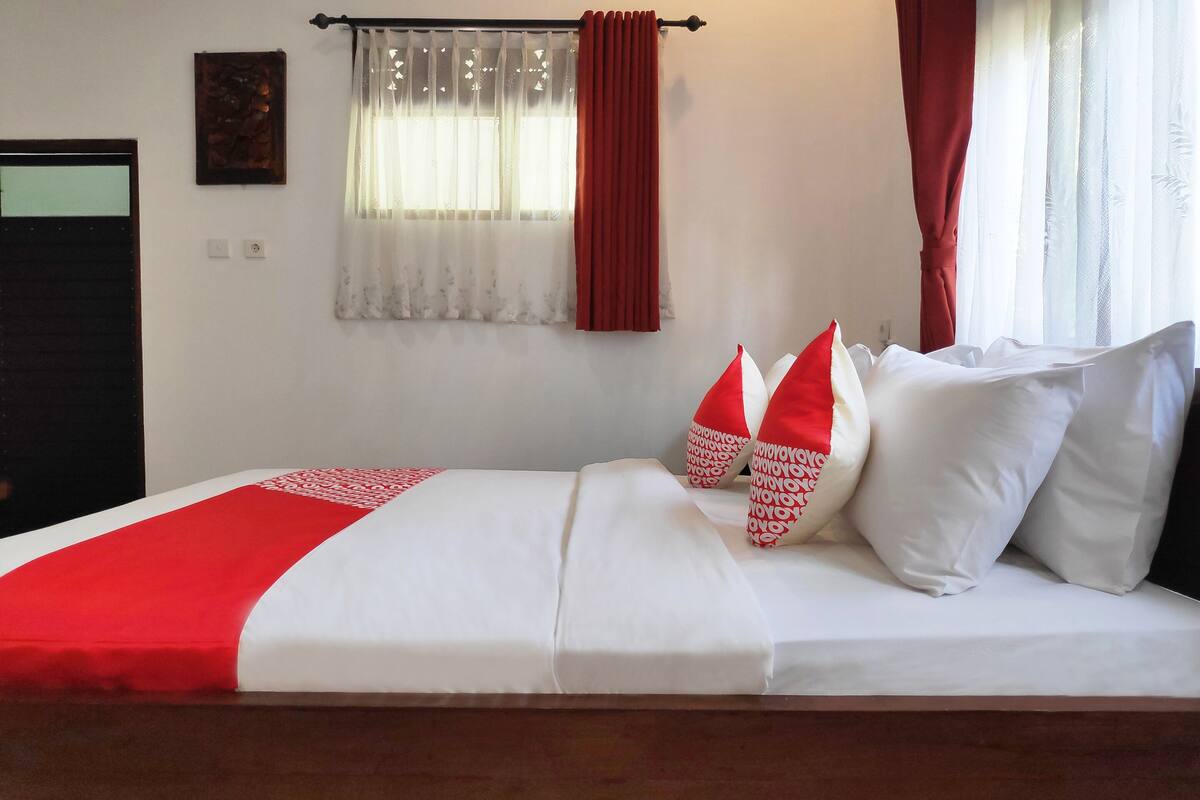 A neatly made bed with white and red bedding, a wooden headboard, and a window with sheer curtains and a red valance.