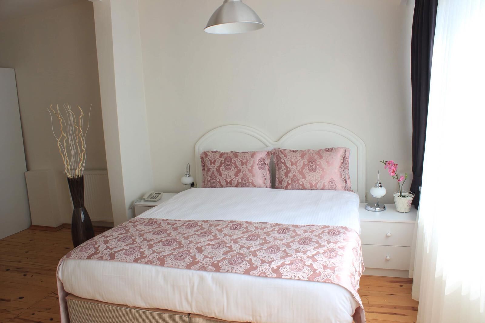Quarto bem arrumado com cama, mesas de cabeceira, abajur e um vaso com flores.