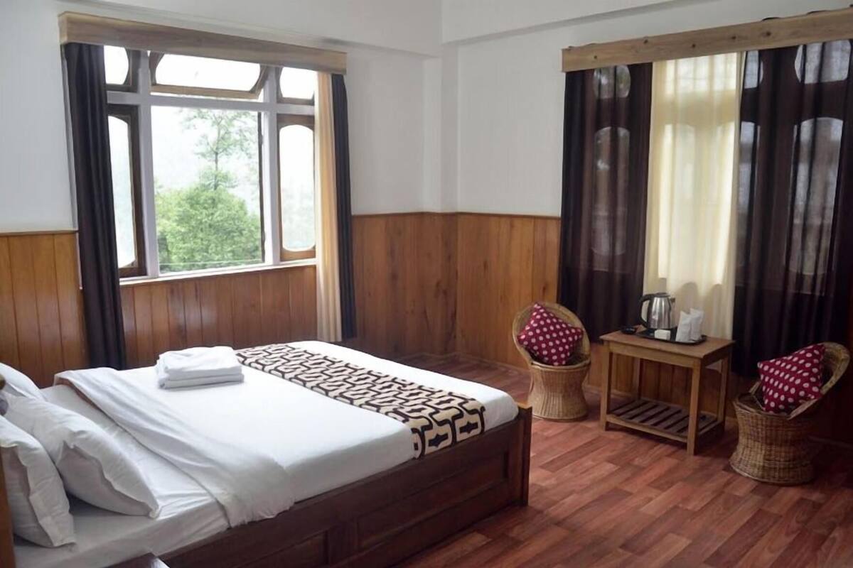 A room with a large bed, a small table with a kettle, two wicker chairs, and a window with curtains.