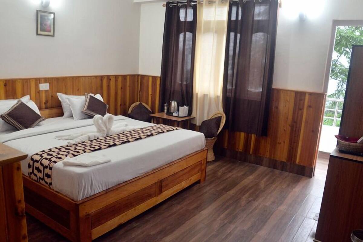 A double bed with a patterned bedspread and pillows, a wooden headboard, a small bedside table with a lamp, and a window with curtains.