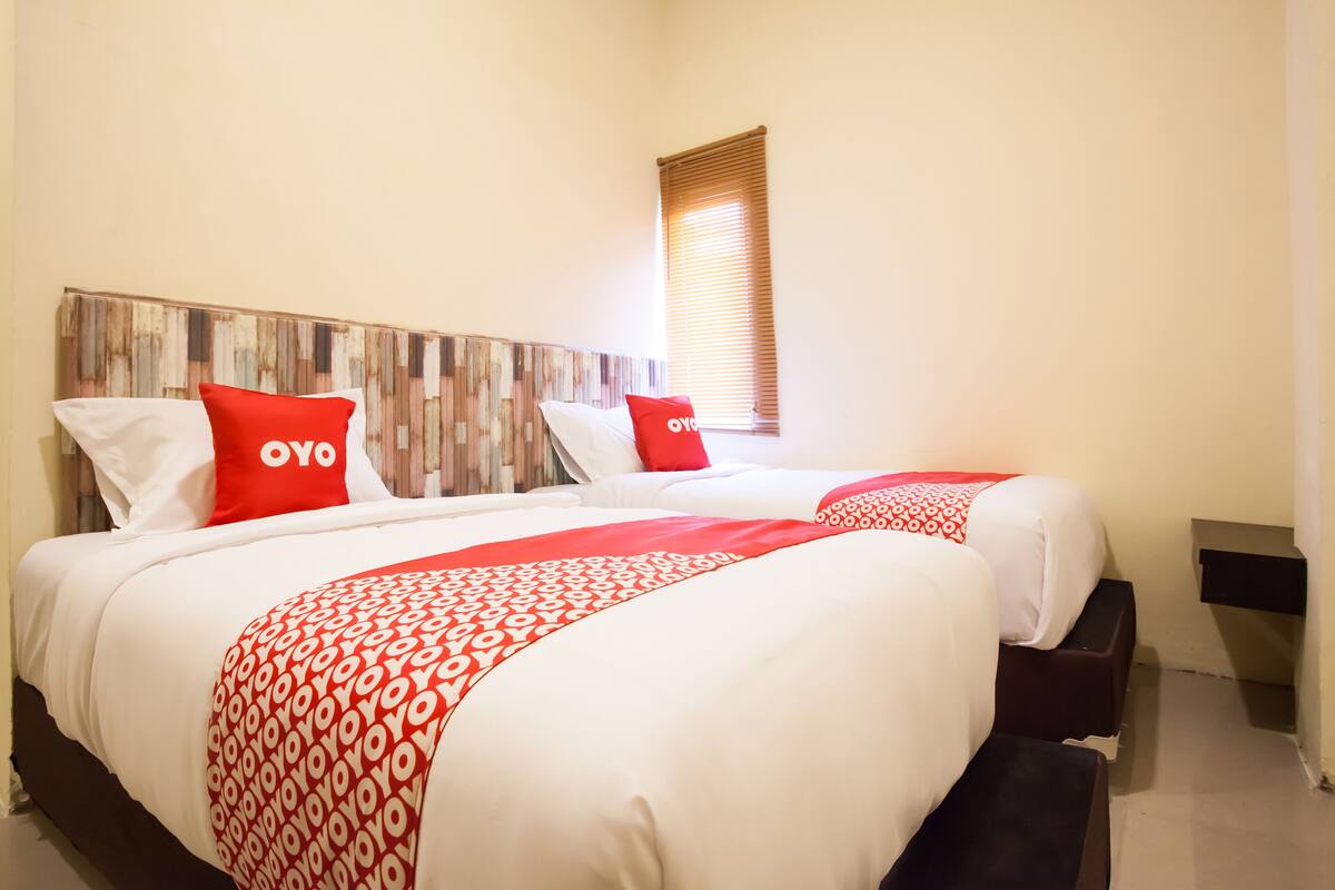 A double bed with white and red bedding, a wooden headboard, and a window with blinds.