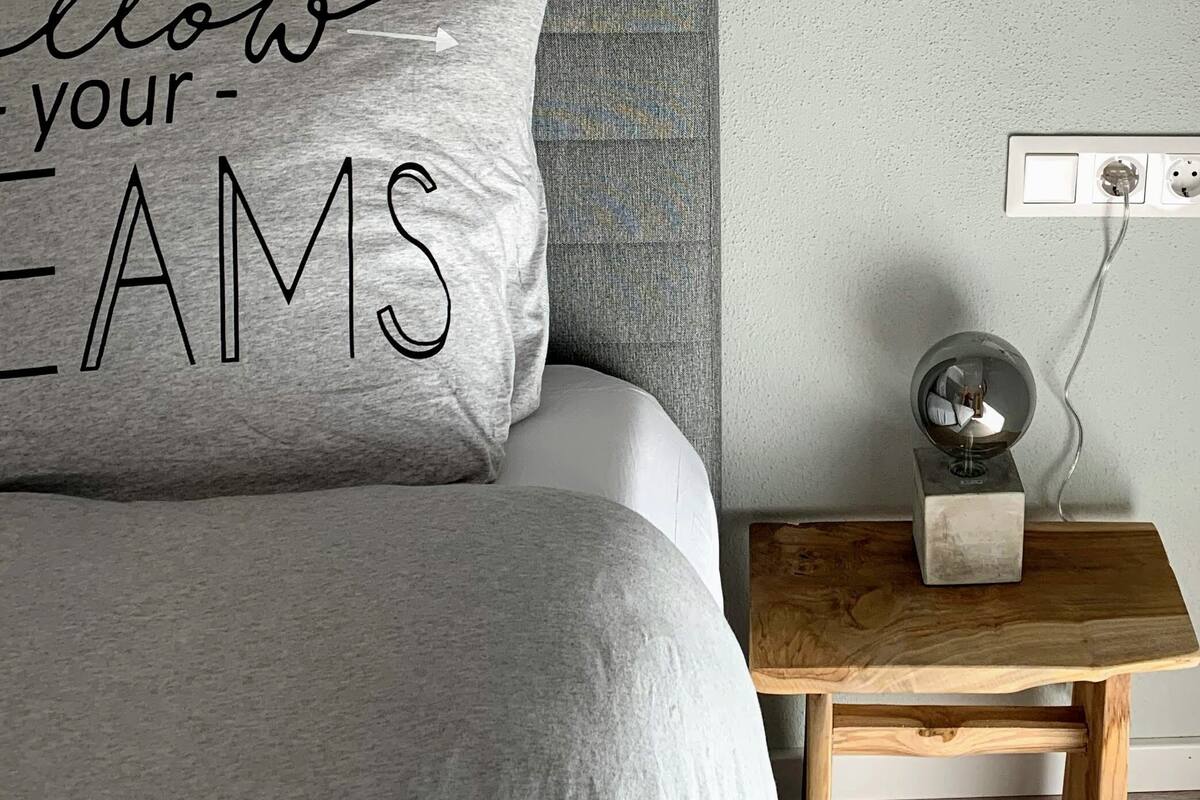 Quarto com cabeceira cinza, cama com edredom cinza e branco, mesa de cabeceira com abajur e uma tomada na parede.