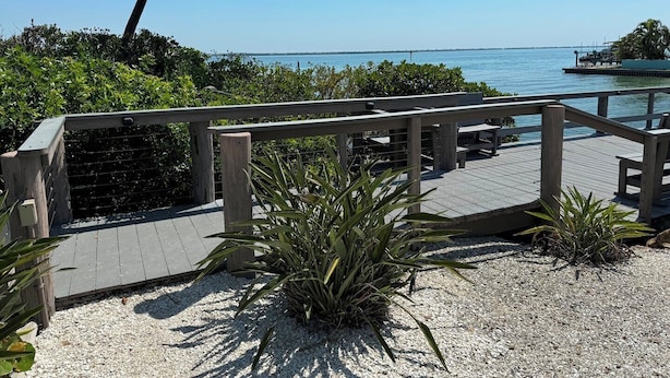 A wooden deck with a railing overlooking a body of water, surrounded by greenery and a clear sky. - Weekly / Monthly Rental - Spacious Condo IN Longboat Key Resort Close TO Pool, Tennis And Beach! (Longboat Key)