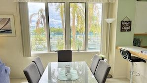 A dining area with a glass table, four chairs, a lamp, and a view of palm trees through large windows. - Weekly / Monthly Rental - Spacious Condo IN Longboat Key Resort Close TO Pool, Tennis And Beach! (Longboat Key)
