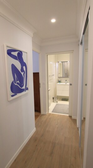 Un couloir doté d’un plancher en bois, une œuvre d’art abstraite encadrée accrochée au mur, et une salle de bain visible à travers une porte ouverte.