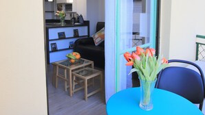 A balcony with a blue table and chairs, a vase with flowers, and a view into a living room with a sofa and a coffee table. - Libération (Nice)